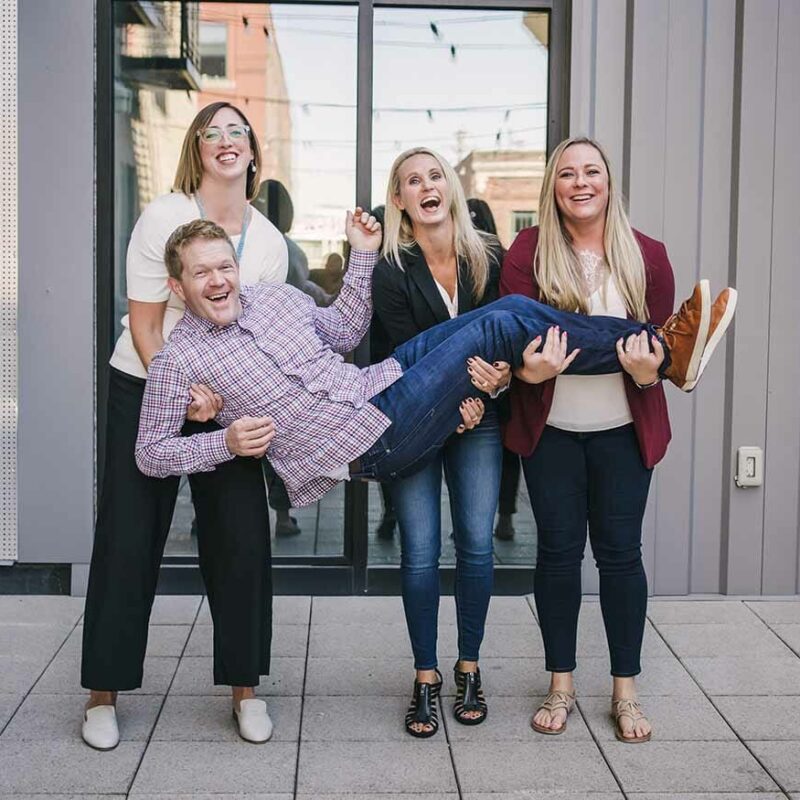 three social assurance employees holding another employee and laughing